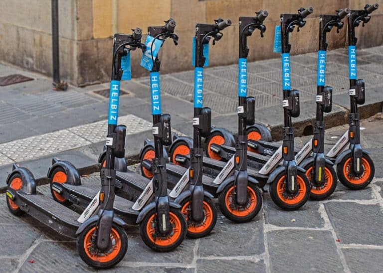 Mehrere Leih-E-Scooter mit orangefarbenen Rädern stehen in einer Reihe auf einem gepflasterten Gehsteig