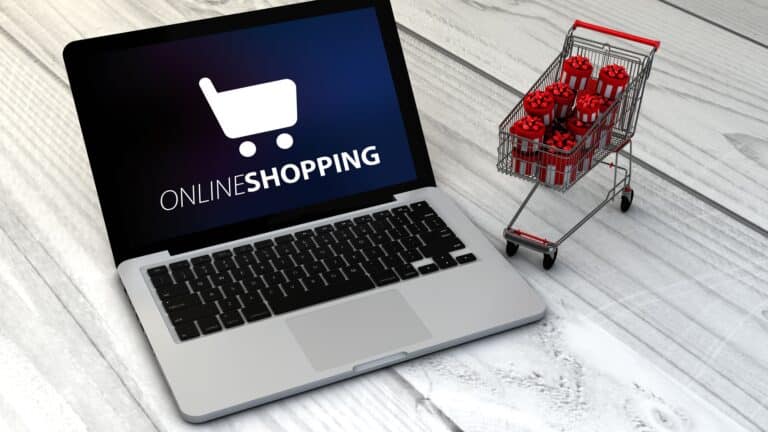 Aufgeklappter Laptop mit dem Schriftzug Online Shopping neben einem kleinen Einkaufswagen voller Geschenkpakete