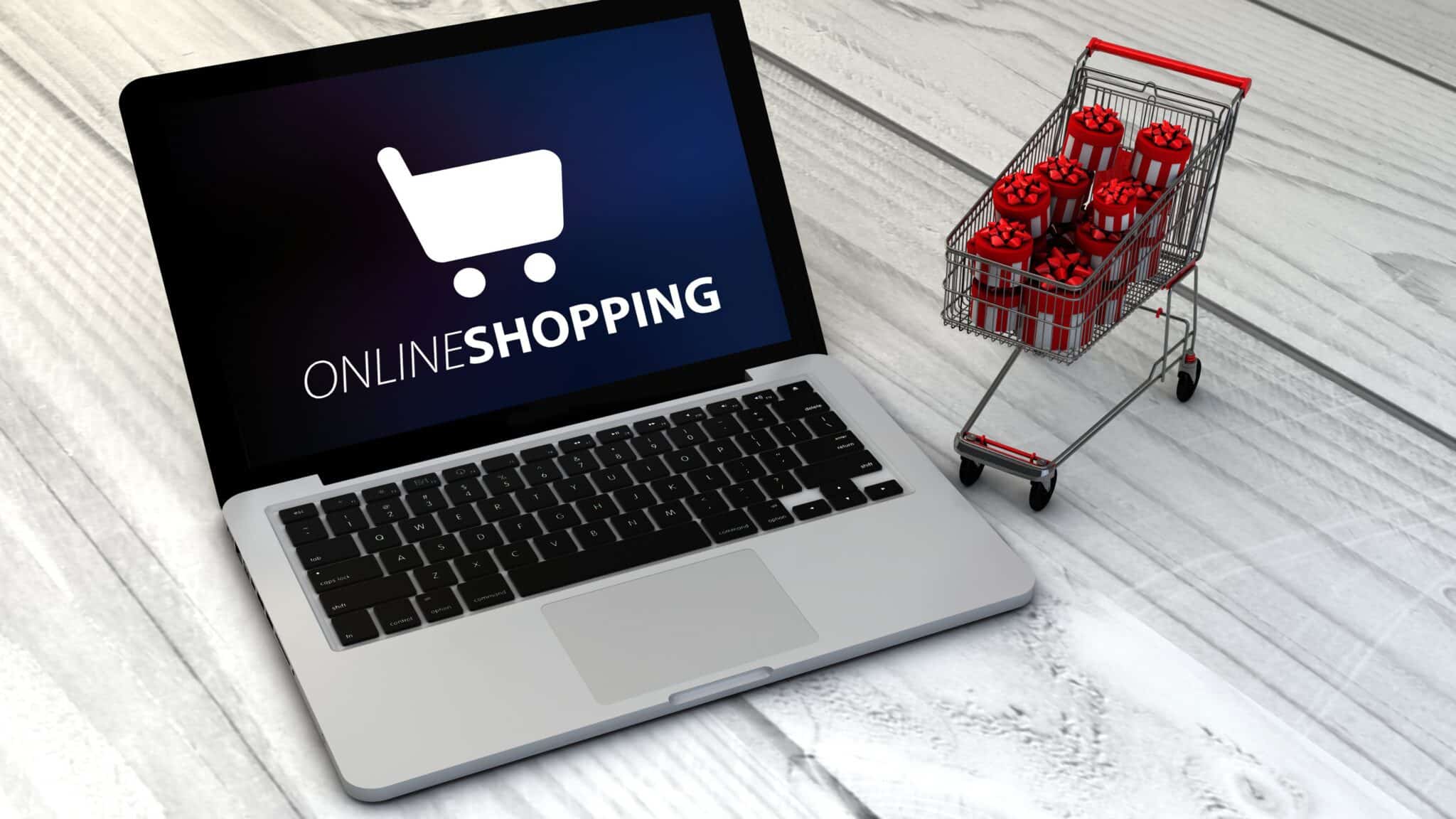 Aufgeklappter Laptop mit dem Schriftzug Online Shopping neben einem kleinen Einkaufswagen voller Geschenkpakete