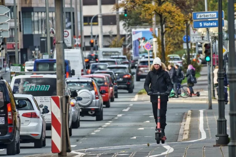 Mann ohne Helm fährt mit einem E-Scooter auf einer Stadtstraße neben einer Autokolonne