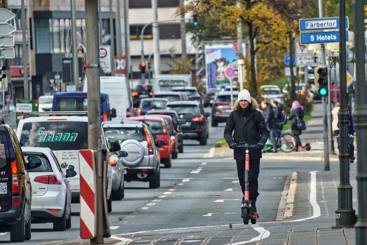 Mann ohne Helm fährt mit einem E-Scooter auf einer Stadtstraße neben einer Autokolonne