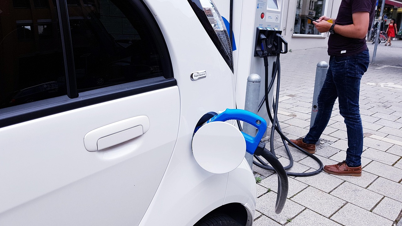 Weißes Elektroauto mit angestecktem Ladekabel an einer Ladesäule, daneben eine Person mit Smartphone